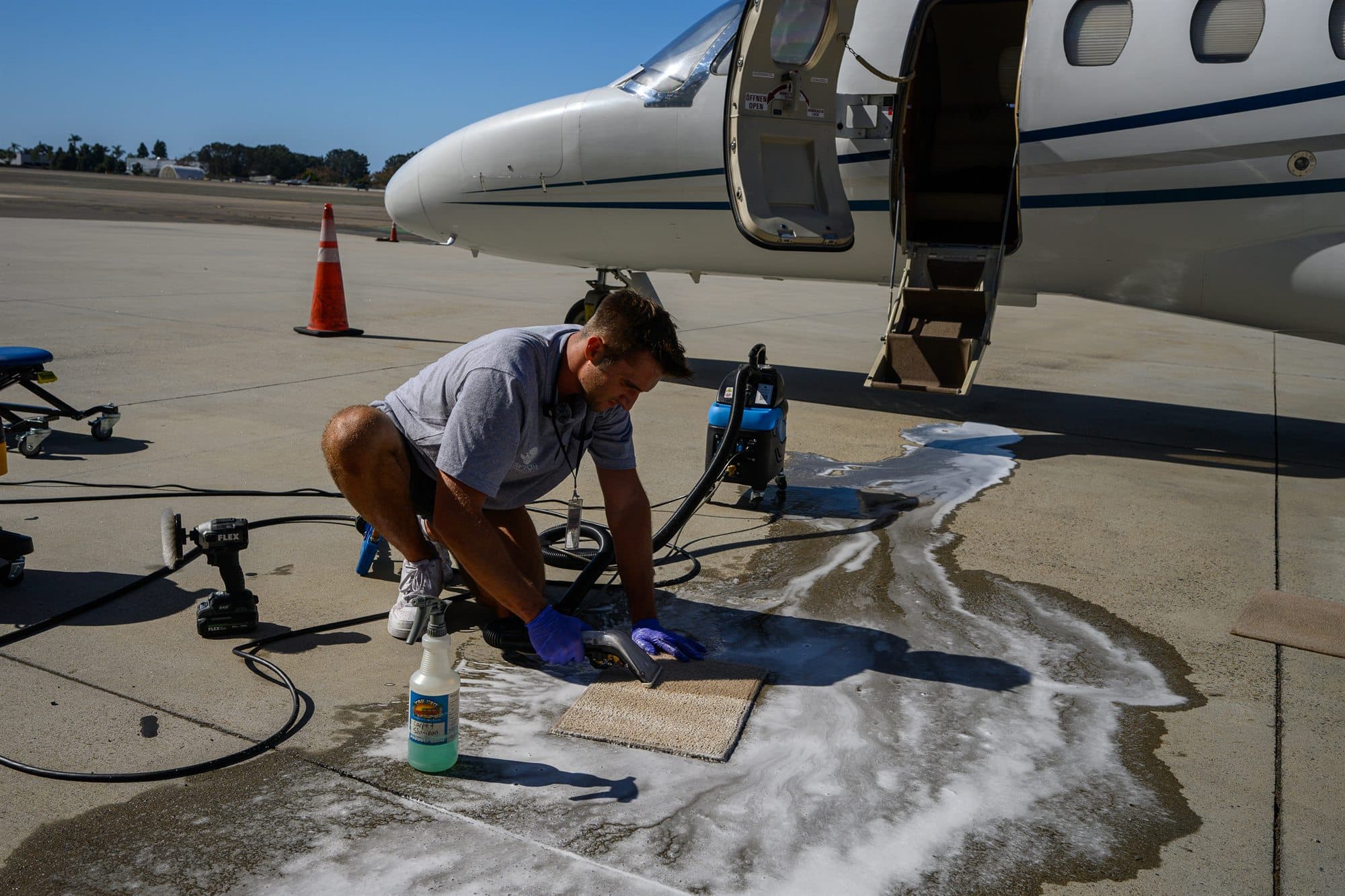 A detailer cleans a cabin carpet panel with an extractor on the ramp beside the aircraft stairs